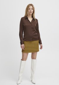 Braunes Satin-Hemd mit Knöpfen, Minirock aus gelb-schwarz kariertem Stoff und weiße kniehohe Stiefel. Glatte Texturen und klare Linien.