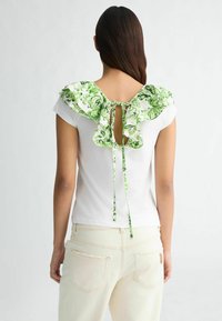 T-shirt blanc avec un col à volants floral vert et un détail de nœud dans le dos. Tissu doux, manches courtes et design avec une ouverture en forme de clé à l'arrière.