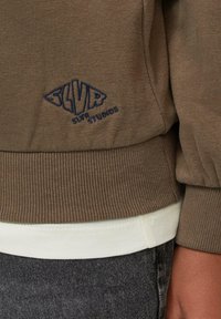 Bruine sweatshirt met geribbelde zoom, voorzien van een blauw geborduurd logo "SLVR STUDIOS." Gelaagd over een wit shirt, gecombineerd met donkere denim.