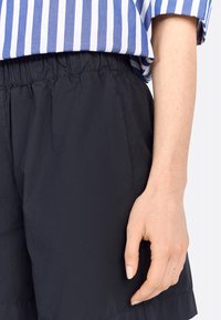 Shorts noirs en tissu léger avec une taille élastique, associés à une chemise rayée bleue et blanche. Main visible reposant sur les shorts.