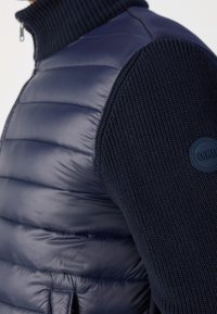 Tummansininen puffervesti, jossa on ribbineulottu hiha ja korkea kaulus. Varustettu pyöreällä logopatchilla hihassa ja pehmustetulla yksityiskohdalla rungossa.