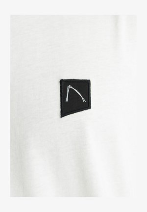 T-shirt bianco in tessuto con una toppa nera ricamata che presenta un design geometrico bianco. Texture liscia, scollatura tonda, maniche corte.