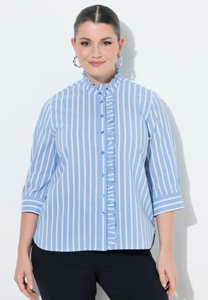 Kobieta z zaczesanymi do tyłu włosami, ubrana w niebiesko-białą paskowaną koszulę z guzikami i falbaną oraz kolczyki koła, stojąca na białym tle.