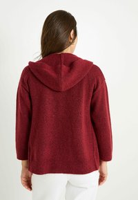 Haut à capuche rouge en tricot avec une coupe décontractée, présentant un tissu texturé, des manches longues et une capuche spacieuse. La vue arrière montre des côtés fendus.