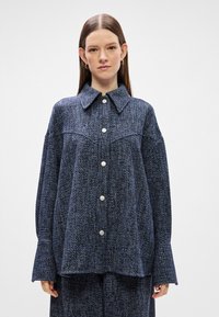 Camisa azul texturizada com colarinho largo, mangas longas e punhos largos, apresentando botões prateados na frente.