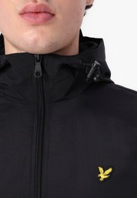 Schwarze wasserdichte Jacke mit Reißverschluss, verstellbarer Kapuze und einem gelben gestickten Adler-Logo auf der Brust. Glatte Textur.