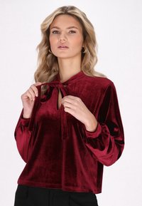 Bordo baršunasta bluza s dekolteom u obliku ključa i detaljem vezanja. Ima dugačke, napuhnute rukave i mekan, teksturiran završetak.