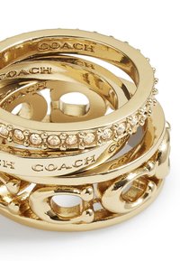 Pile de bagues Coach en or, l'une ornée de cristaux incrustés et les autres gravées du nom de la marque, montrées en gros plan sur fond blanc.
