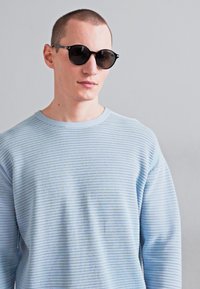 Pulover tricotat de culoare albastru deschis, cu un model texturat, guler rotund și mâneci lungi. Ochelari de soare rotunzi, negri, cu lentile întunecate.