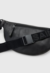 Zwarte synthetische leren schoudertas met een gladde textuur, een voorvak met rits en een brede verstelbare band met het woord "SAINT" in vetgedrukt.