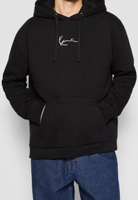 Man som bär en svart hoodie med ficka fram och vit "Kani" logotyp skriven i kursiv, tillsammans med blå jeans. Händerna i hoodie-fickan.