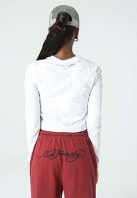 Pitkähihainen valkoinen paita, jossa on hienovarainen graafinen kuvio, yhdistettynä viininpunaisiin verkkareihin, joiden takana on musta "Ed Hardy" -teksti. Harmaa lippalakki päässä.
