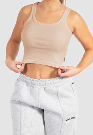 Kvinde iført beige crop top og lysegrå joggingbukser, der holder i kanten af toppen med begge hænder mod en ensfarvet baggrund.
