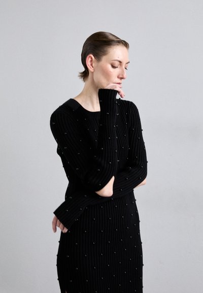 Abito nero a costine con maniche lunghe, caratterizzato da piccole decorazioni rotonde e scollatura a barca. Il design è aderente con spacchi laterali.