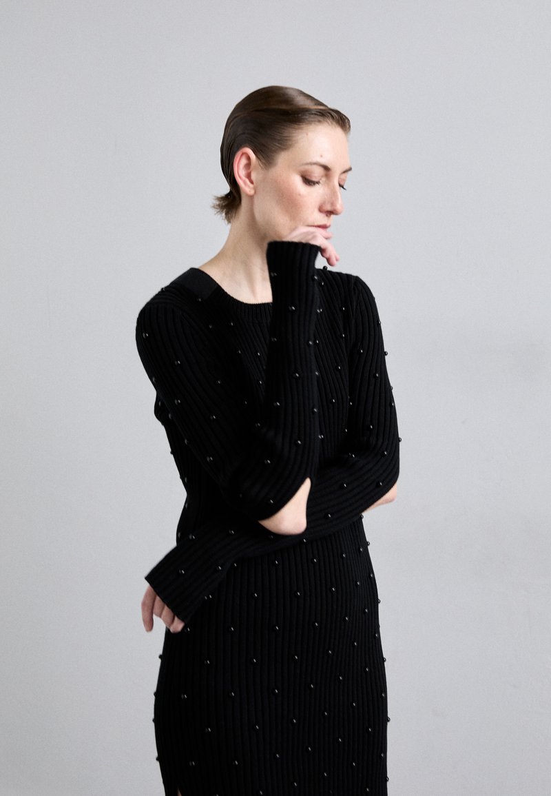 Robe noire côtelée à manches longues, ornée de petites décorations rondes et d'un décolleté bateau. La coupe est ajustée avec des fentes sur les côtés.