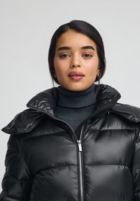 Svart dunjacka med glansig yta, utrustad med hög krage, dragkedja framtill och strukturerade sömmar för värme och komfort.