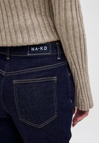 Mörkblå denimjeans med gula sömmar och två bakfickor; kombinerade med en ljusbeige ribbad tröja. NA-KD-märket synligt.