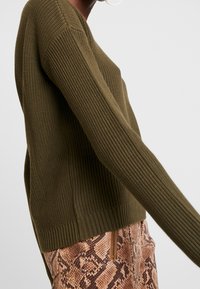 Oliivinvihreä, resorineuloksella varustettu neulepusero, jossa pitkät hihat ja vartalonmyötäinen helma, yhdistettynä ruskeisiin, käärmekuvioituun tekstuurihousuihin.