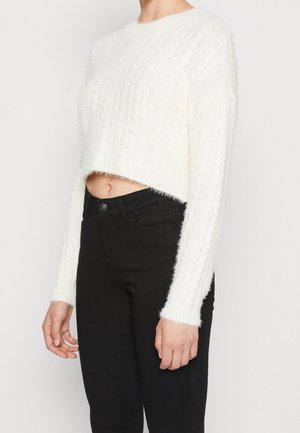 Kvinde iført en hvid, blød, cropped sweater med lange ærmer og højtaljede sorte bukser, vist fra hals til hofter.