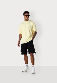 Hellgelbes, oversized T-Shirt mit kleinem Logo in der Mitte, kombiniert mit schwarzen Latzhosen mit Seitentaschen und weißen Sneakers.