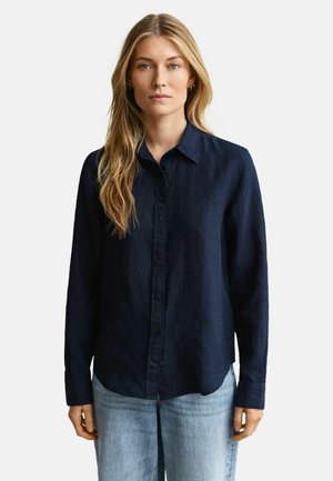 Femme aux longs cheveux blonds portant une chemise boutonnée bleu marine et un jean bleu clair, debout devant un fond blanc uni.
