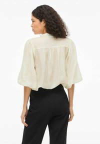 Blusa de color beige claro con tejido texturizado y transparente, mangas abullonadas, y un detalle de yugo en la parte trasera, combinada con pantalones negros de tiro alto.