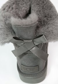 Botte en suède gris avec une doublure en peluche grise, forme arrondie et un nœud décoratif en ruban gris à l'arrière. Logo UGG visible.