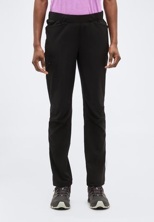 Vue de face d'une personne portant un pantalon noir à taille élastique avec poches latérales et des chaussures de randonnée grises sur un fond clair uni.