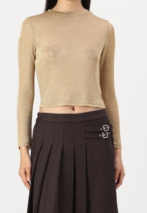 Femme portant un crop top doré à manches longues associé à une jupe plissée marron foncé taille haute avec deux boucles latérales.