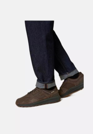 Person trägt dunkelblaue hochgekrempelte Jeans und braune Freizeitsneaker mit schwarzen Socken vor einem schlichten weißen Hintergrund.