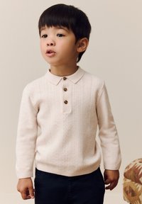 Maglione rosa chiaro a trecce con colletto e tre bottoni marroni, abbinato a pantaloni blu scuro, indossato da un bambino con capelli scuri.