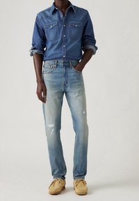 Tmavě modrá džínová košile se dvěma předními kapsami a bílými knoflíky, doplněná světle modrými roztrhanými džínami a béžovými mokasínami.