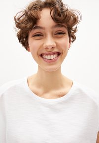 Witte, kortebloemige t-shirt met een zachte textuur en een ronde halslijn, gedragen door een persoon met kort, krullend bruin haar, die glimlacht tegen een lichte achtergrond.
