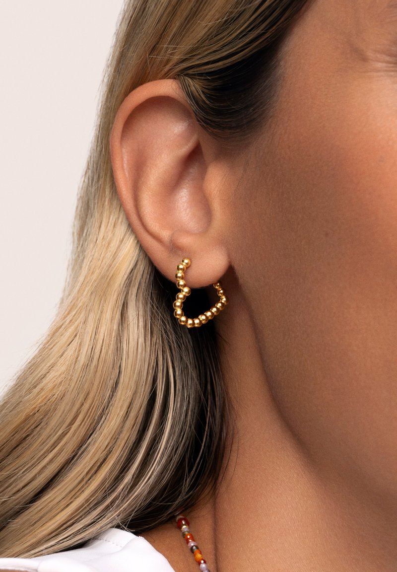 Pendientes de aro de oro con un diseño texturizado, presentando pequeñas cuentas redondeadas que forman una forma ondulada. Llevados en una oreja con cabello rubio largo.