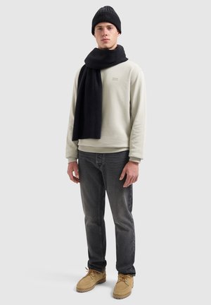 Lichtgrijze sweatshirt, zwarte gebreide sjaal en beanie, gecombineerd met donkere jeans en beige schoeisel. Geperforeerde materialen zijn overal zichtbaar.