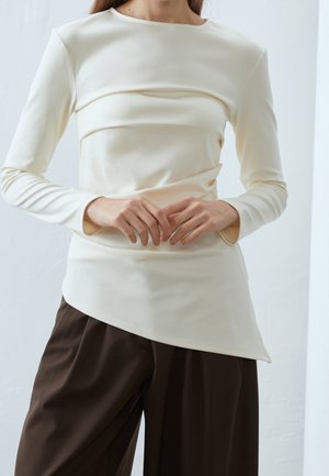 Femme portant un haut à manches longues écru avec un ourlet asymétrique et un pantalon large marron foncé, debout, les mains légèrement jointes.