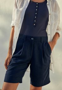 Granatowe szorty z zakładkami i bocznymi kieszeniami, wykonane z lekkiego materiału. Połączone z ciemnym prążkowanym topem i białą teksturowaną koszulą.