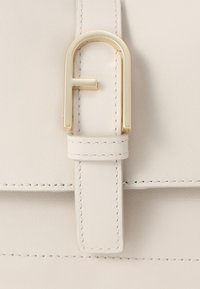 Λευκή δερμάτινη τσάντα με αγκράφα που διαθέτει χρυσό μεταλλικό σχέδιο σε σχήμα 'F'. Λεπτή ραφή ορατή στο λουρί και στην επιφάνεια της τσάντας.