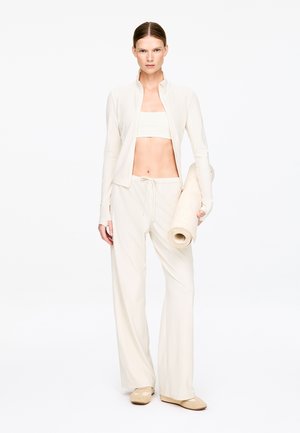 Jasnobeżowy sportowy strój składający się z dopasowanego crop topu i luźnych spodni, wykonany z miękkiego, elastycznego materiału, z długimi rękawami oraz z rolowaną matą do jogi.