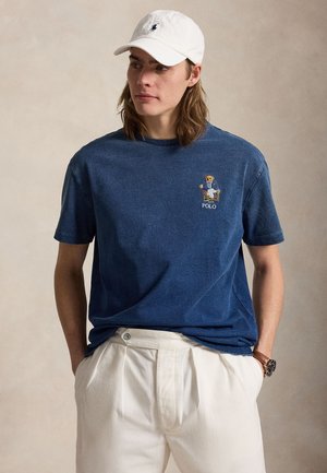 Jonge man met lang haar die een witte pet draagt, een blauwe polo met beer, witte broek en een polshorloge, staand tegen een effen achtergrond.
