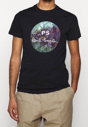 Uomo che indossa una maglietta nera con una grafica circolare colorata e il testo "PS Paul Smith", abbinata a pantaloni beige, con le mani rilassate.