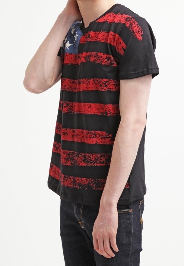 Svart T-shirt med horisontella slitna röda ränder och en blå sektion med vita stjärnor på vänster axel. Korta ärmar, avslappnad passform.