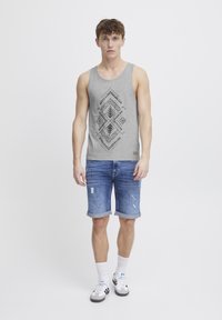 Graues Tanktop mit schwarzem geometrischem Druck, kombiniert mit zerrissenen blauen Jeansshorts. Das Model trägt weiße Socken und graue Sneakers.
