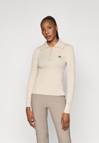Lohkoperho beige ribbipaita pitkillä hihoilla, nappikaulus, tasainen kaulus ja ruskea brodeerattu logo vasemmassa rinnassa. Silkkinen pinta.