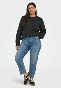 Frau trägt einen dunkelgrauen Strickpullover, hellblaue hochgeschnittene Jeans, einen schwarzen Gürtel und schwarz-weiße Sneaker und steht mit einer Hand in der Tasche.