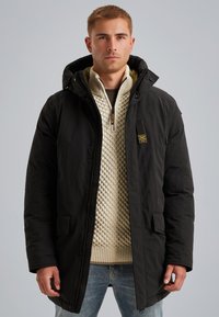 Zwarte gewatteerde parka met een ritssluiting, voorzien van een patch pocket en geribde manchetten. Draagt over een crèmekleurige gestructureerde trui en blauwe denim.