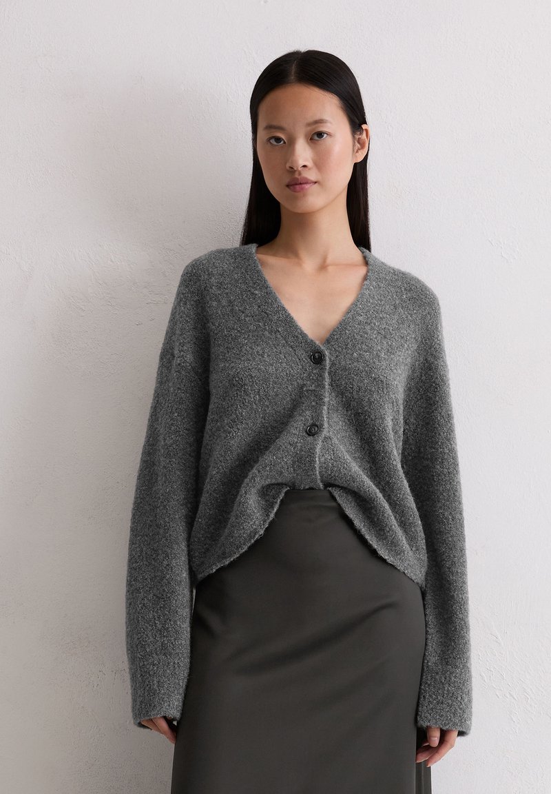 Vrouw met lang zwart haar, gekleed in een grijze fuzzy cardigan met knopen en een donkergrijze rok, staande tegen een effen lichte muur.