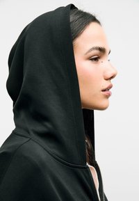 Crna zip-up trenirka s glatkom teksturom i velikom kapuljačom koja pada. Dizajn ima vidljivo šivanje duž šavova.