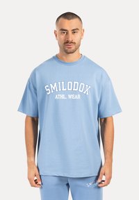 Hellblaues, oversized T-Shirt aus weichem Stoff mit einem weißen "SMILODOX ATHL. WEAR" Logo, das auf der Vorderseite aufgedruckt ist. Kurze Ärmel, Rundhalsausschnitt.