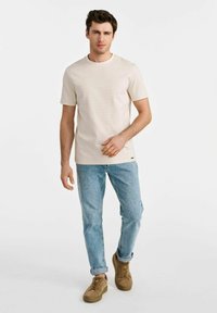 Jasno beżowa koszulka z krótkim rękawem i okrągłym dekoltem, zestawiona z jasnoniebieskimi dżinsami i brązowymi zamszowymi trampkami.